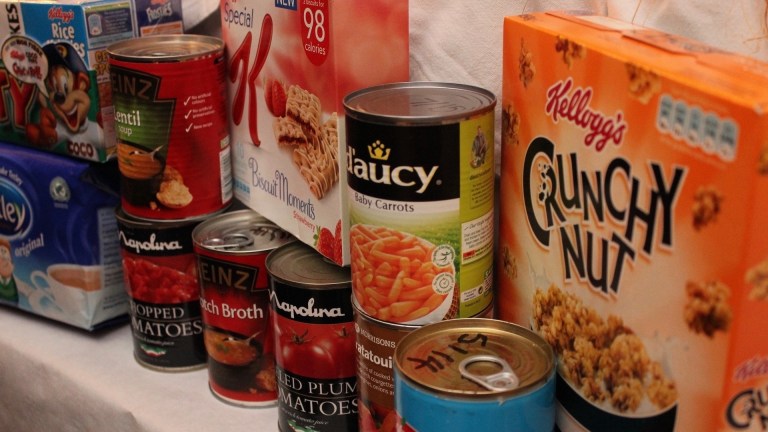 cans and boxes of food sit on a shelf. children in poverty are being offered tiny amounts in parcels sent to replace free school meals