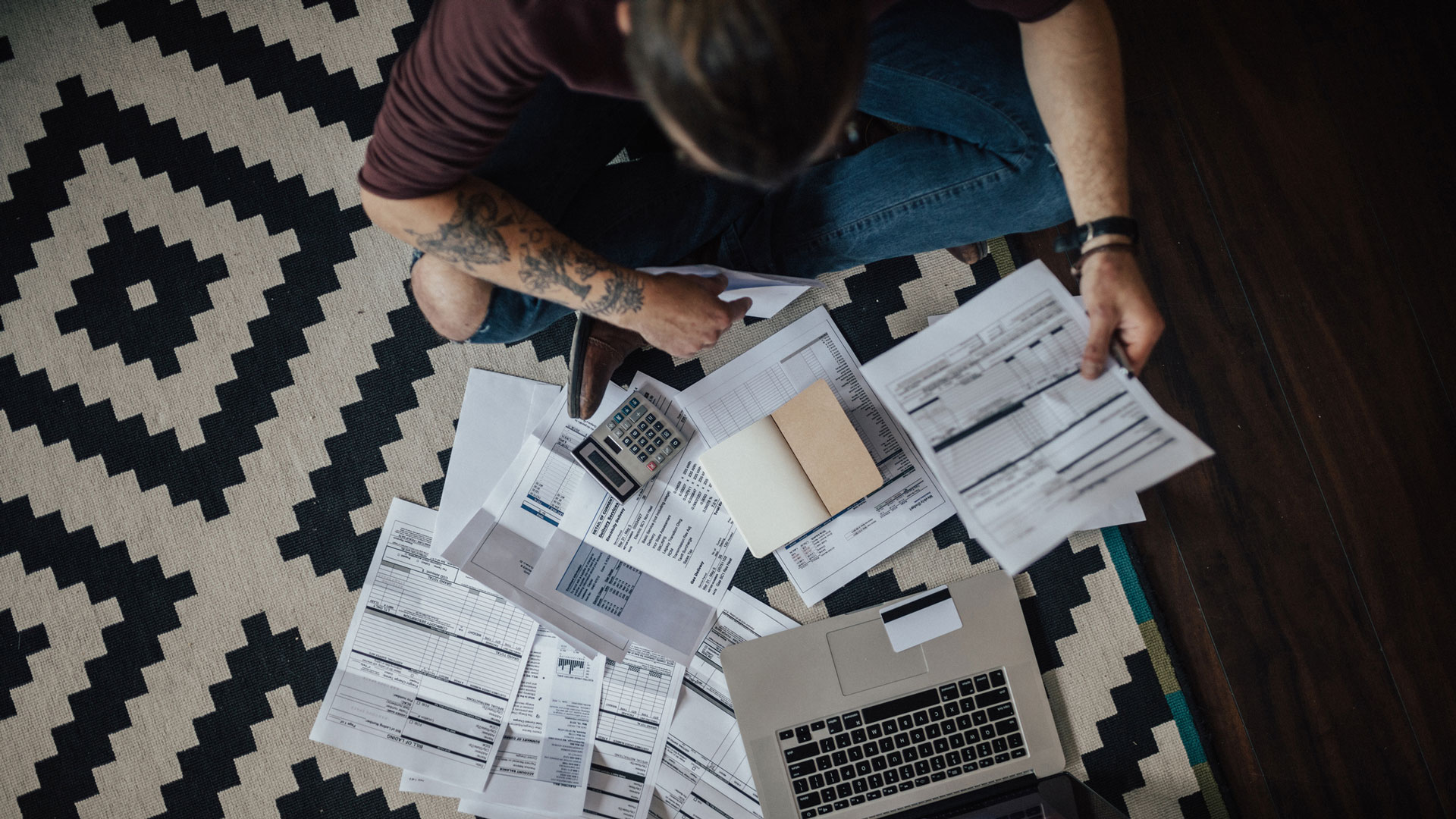 A young person, seen from above, sits on the floor looking through a stack of bills in front of a laptop