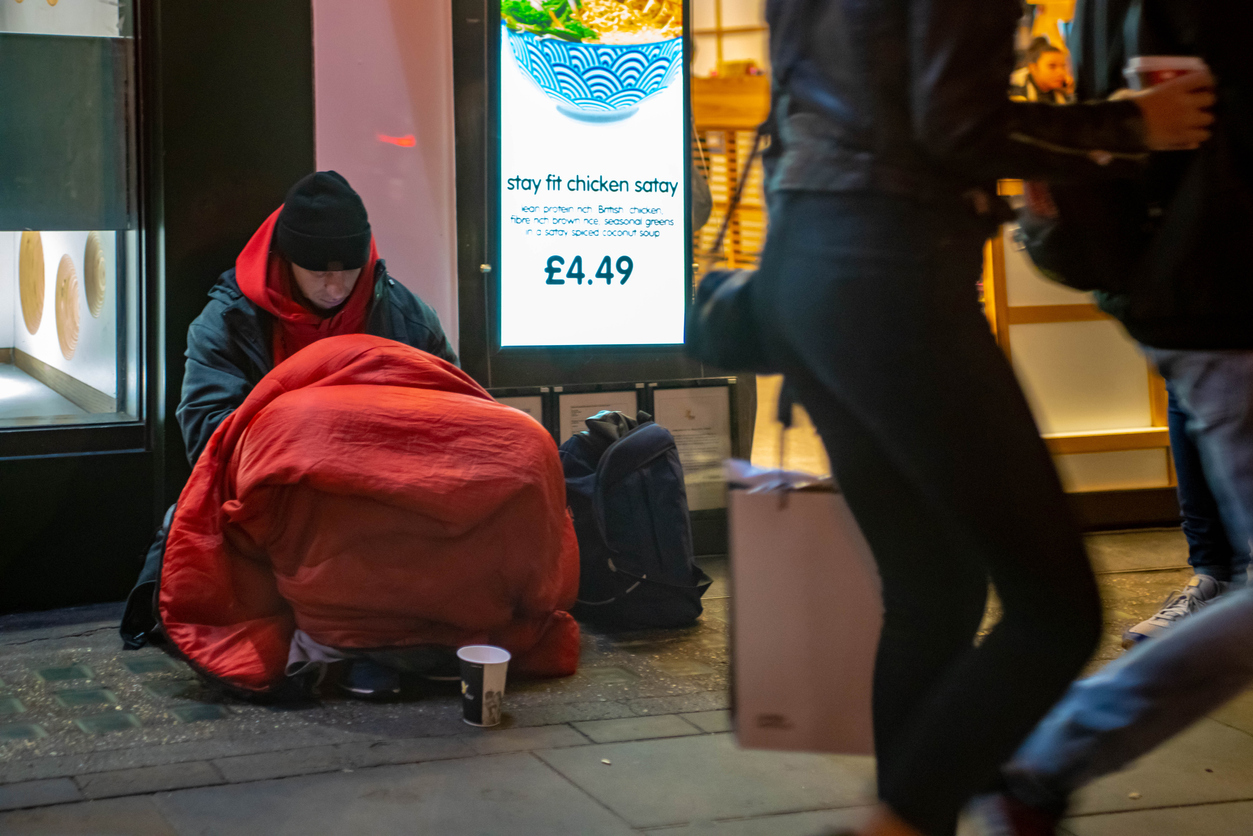 A homeless man sits on the floor of a trin station, wrapped in blankets with a cup in front of them