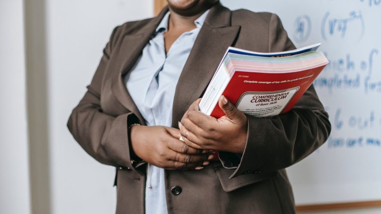 A woman holds books in front of a whiteboard in a classroom