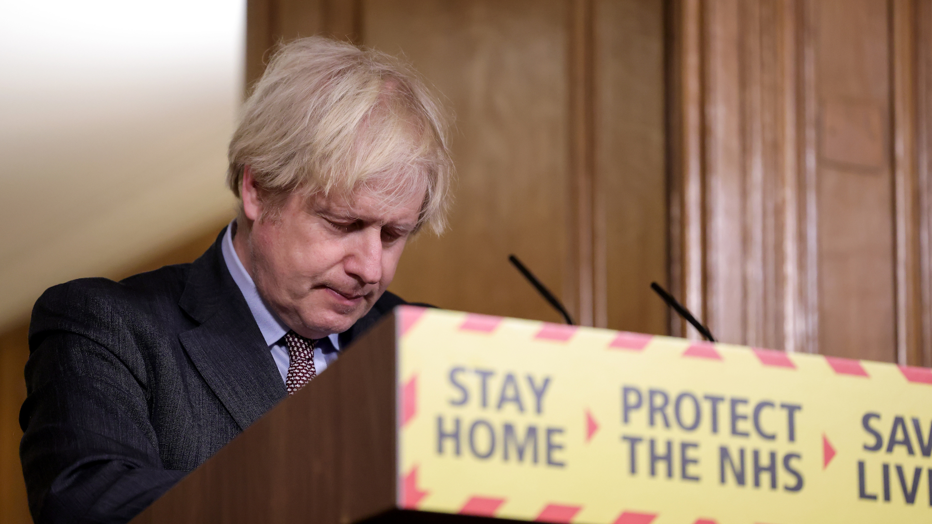 Boris Johnson stands at a podium with government warnings, with his head bowed