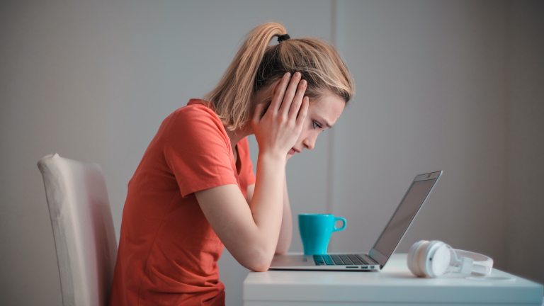 A young woman sits with her head in her hands, looking at a laptop