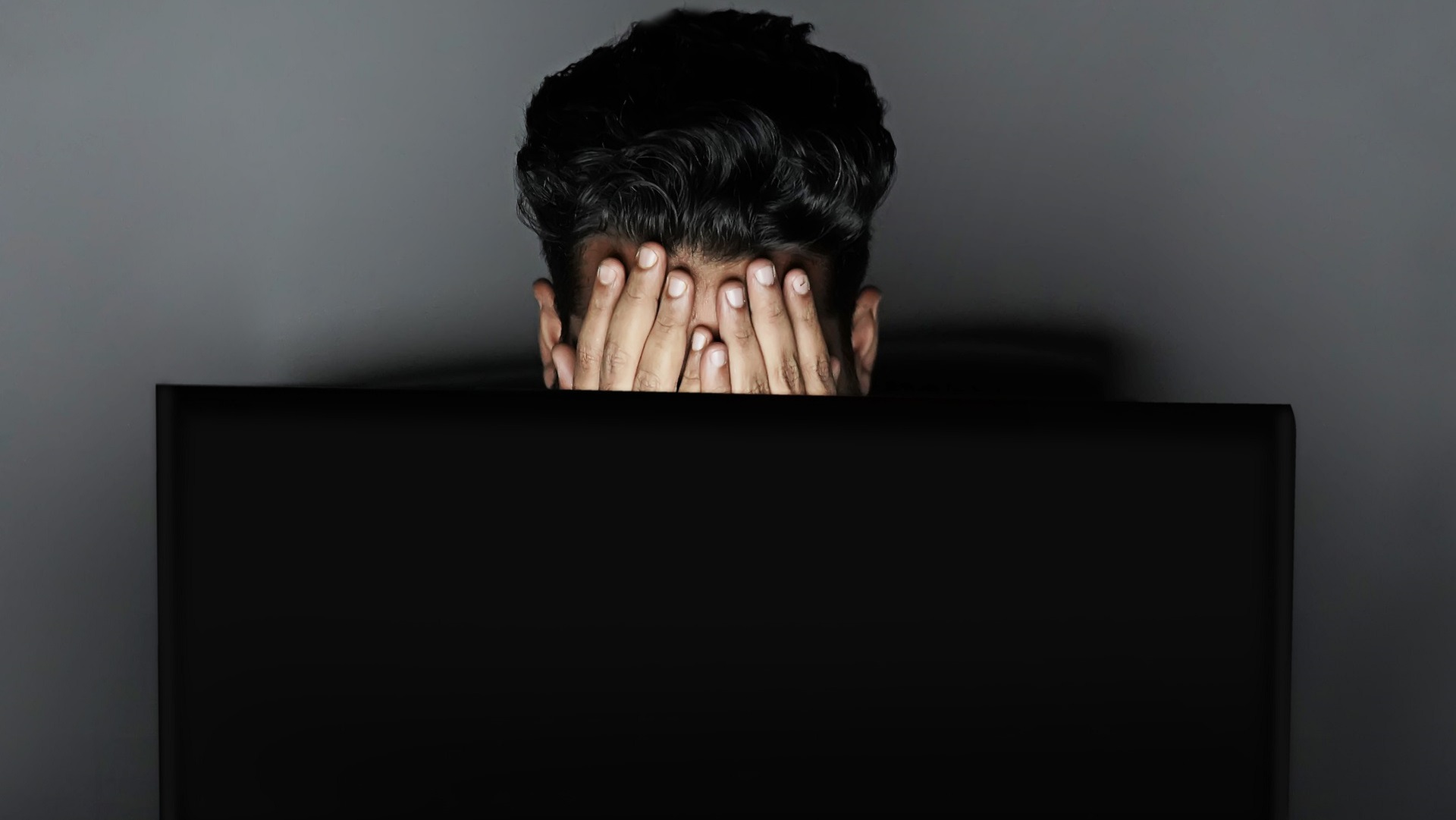 A young man holds his head in his hands at a laptop, worried about unemployment