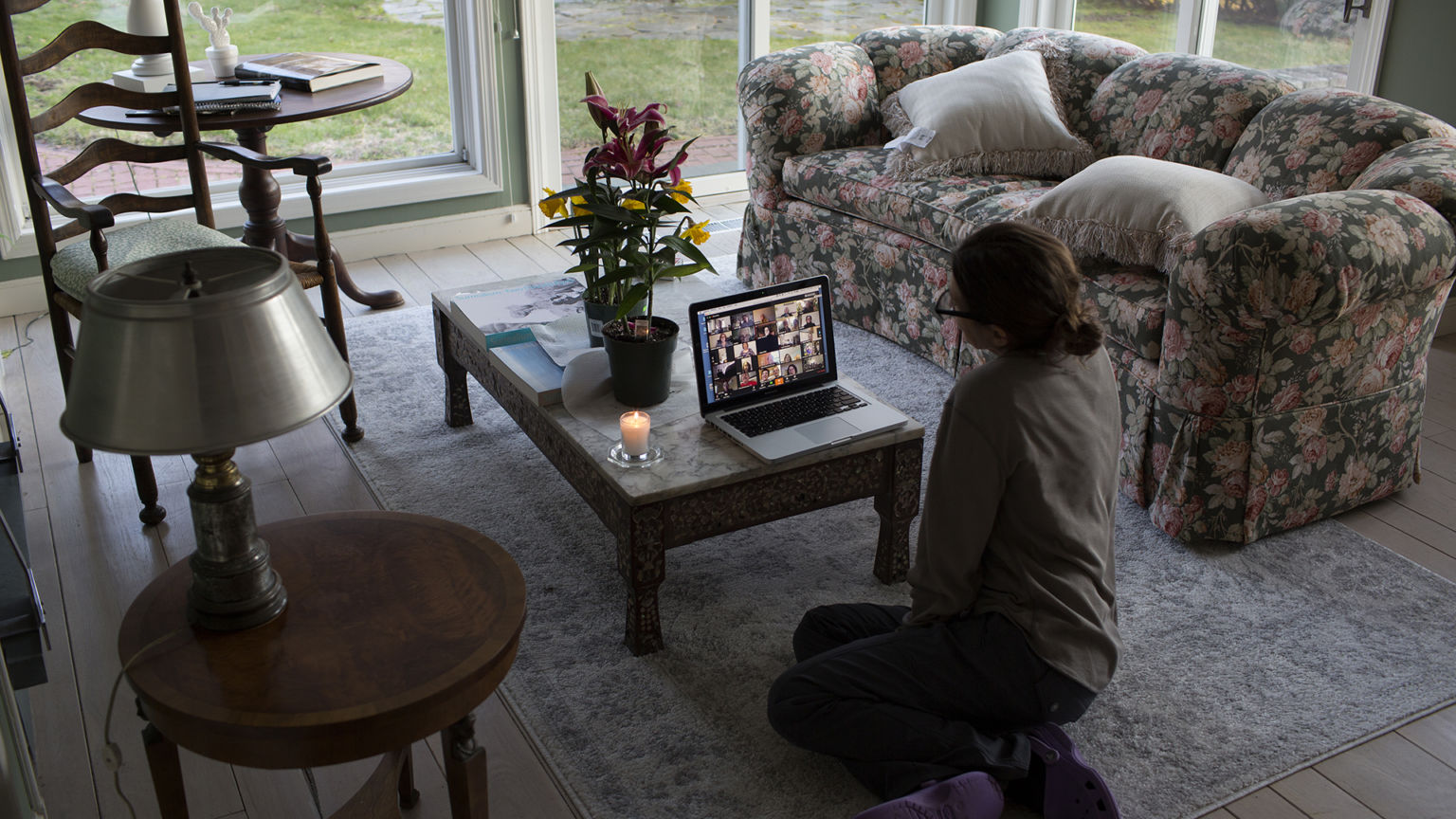 A woman sits alone in a sitting room looking at faces on a digital call, with a single candle alongside.