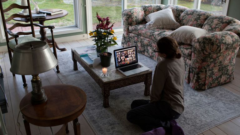 A woman sits alone in a sitting room looking at faces on a digital call, with a single candle alongside.