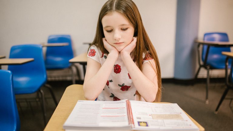 A young girl sits at her school desk staring down at her textbook and looking lonely. Mental health