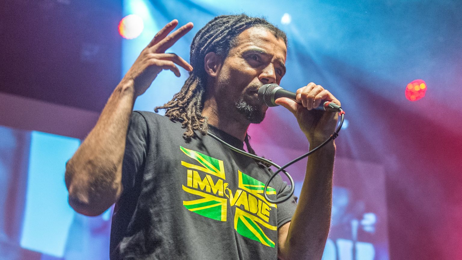 Image shows artist Akala with a microphone in hand, wearing a t-shirt with lights shining behind him