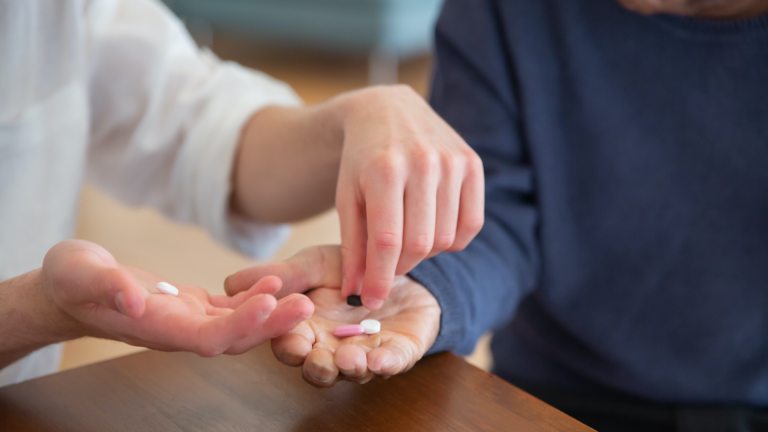 woman's hands placing medication into elderly man's hands. social care cap