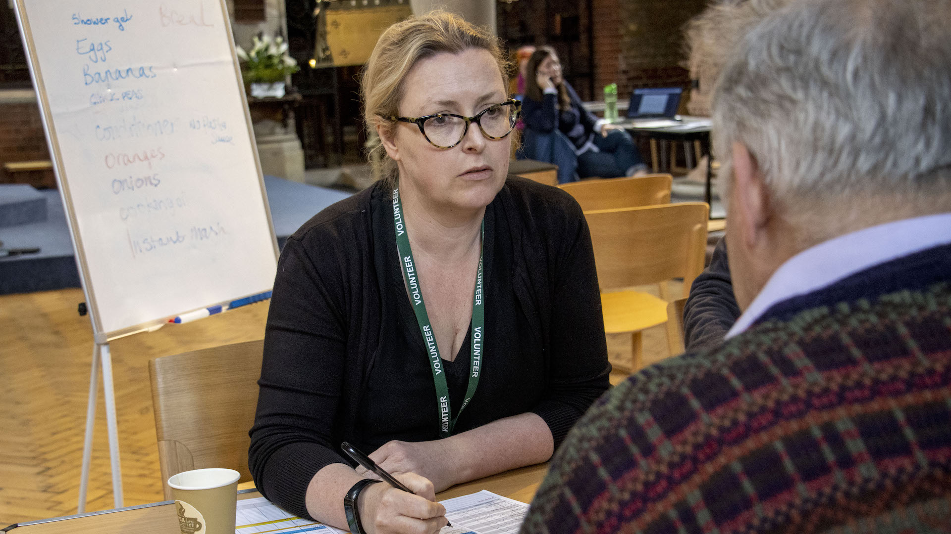 A woman looks concerned and holds a pen, behind her a board lists food items used at Earlsfield Foodbank