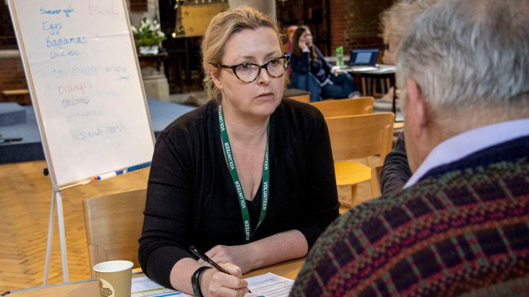 A woman looks concerned and holds a pen, behind her a board lists food items used at Earlsfield Foodbank