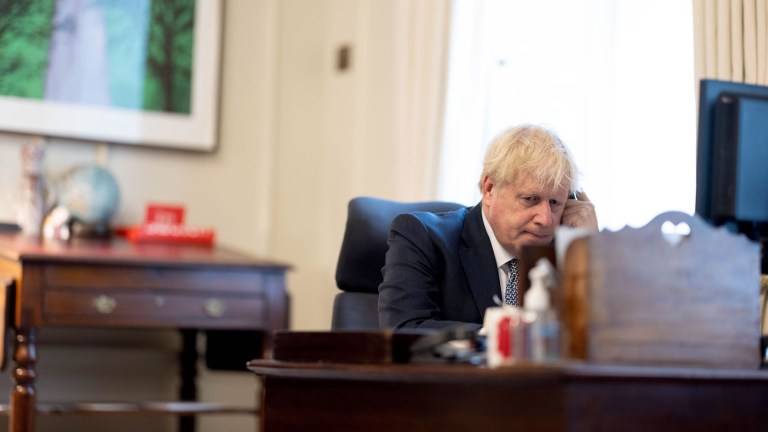 Boris Johnson sits at a dark wood desk with a phone in his hand and serious expression on his face