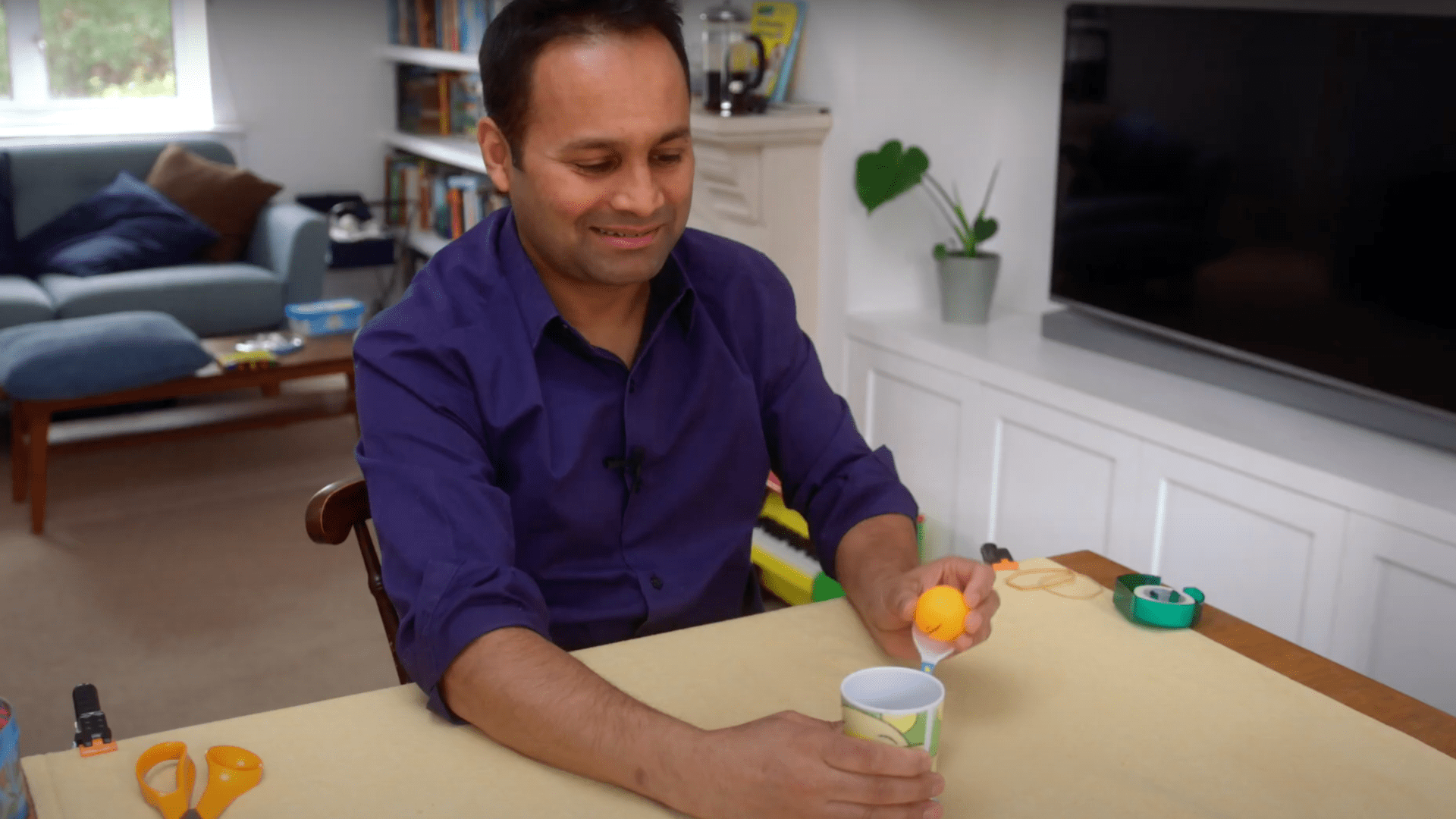 Alom Shaha wears a blue shirt sitting at a table and preparing to firehis mini catapult made from a spoon and cup