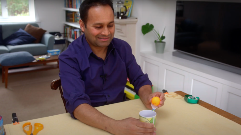 Alom Shaha wears a blue shirt sitting at a table and preparing to firehis mini catapult made from a spoon and cup