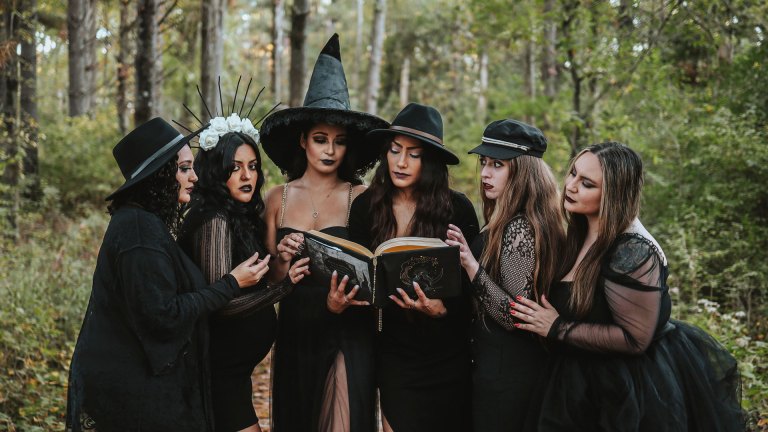 Group of women dressed as witch coven reading spell book in forest.