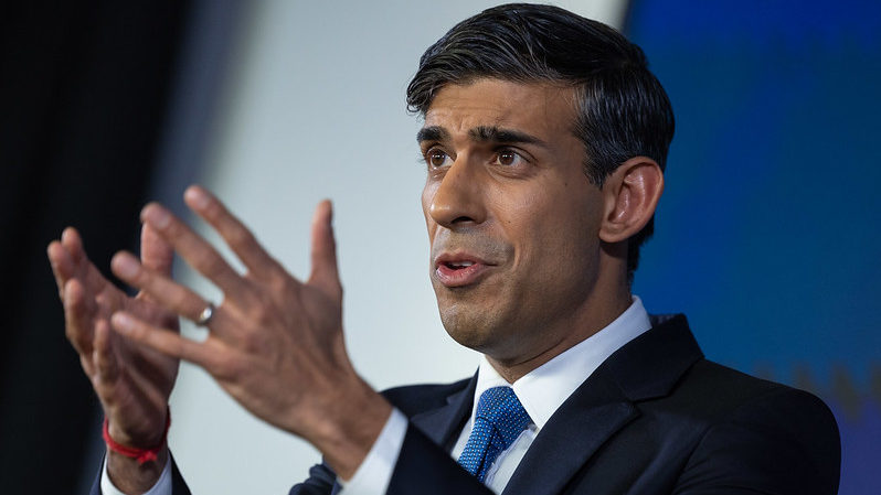 Rishi Sunak stands with his hands in front of him while giving a speech