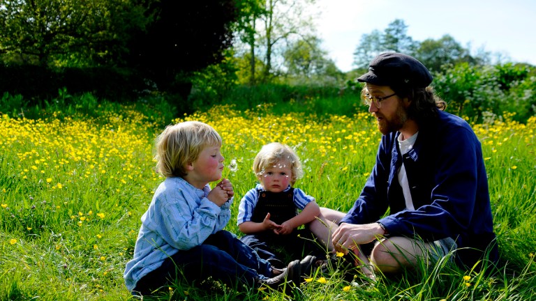 Two red faced children and a man sit in a meadow in summer surrounded by long grass and wildflowers