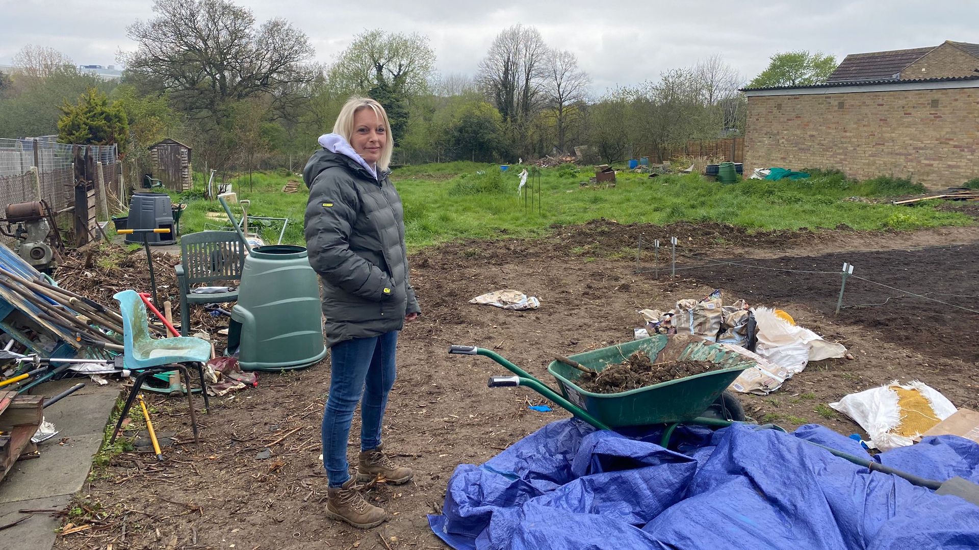 Carly Burd standing in a down jacket, jeans, and boots next to a wheelbarrow full of soil in her allotment