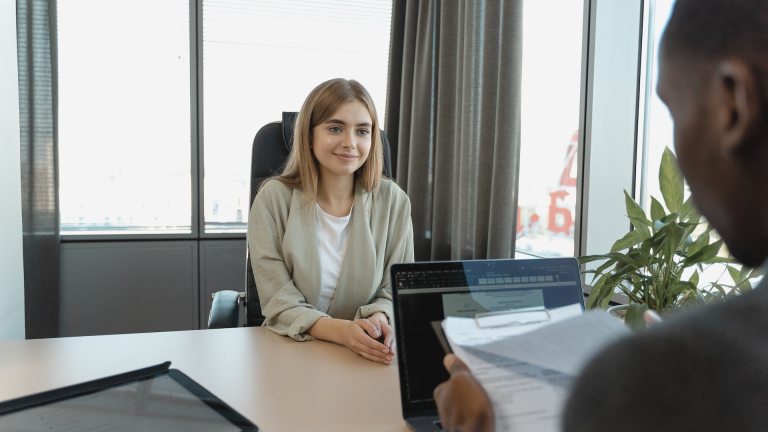 A young woman sits at a desk while a man looks at a note pad and laptop