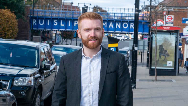 Danny Beales walks down a street in Ruislip Manor with a blue bridge in the background spelling the name of the area