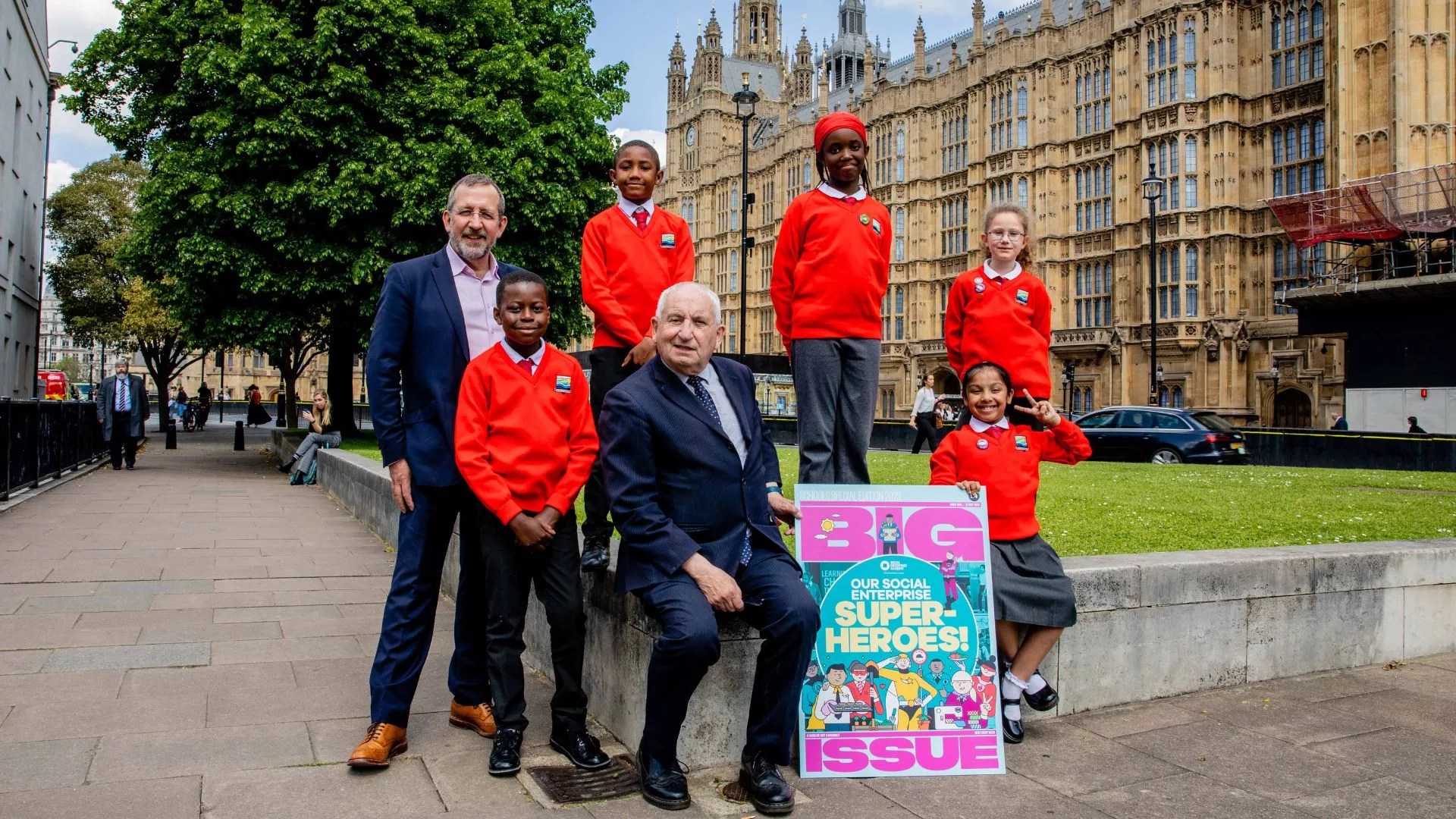 A group of school children in uniform and two white men in dark suits outside the Houses of Parliament in London on a bright, sunny day,