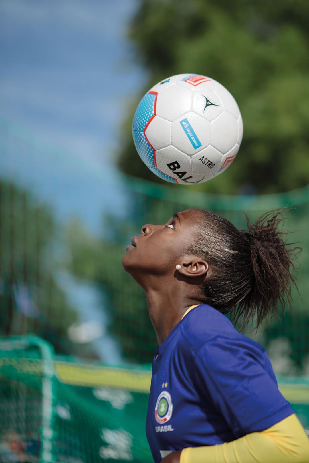 A female Brazilian football player in a dark blue jersey heads the ball