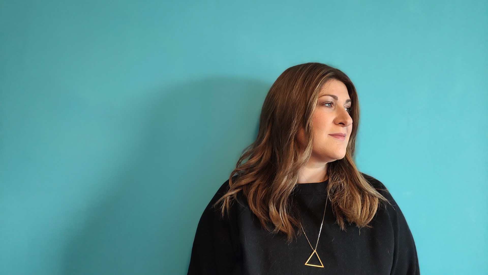 Lara Roberts, a young woman in her thirties with shoulder length brown hair, looks away against a pale blue background