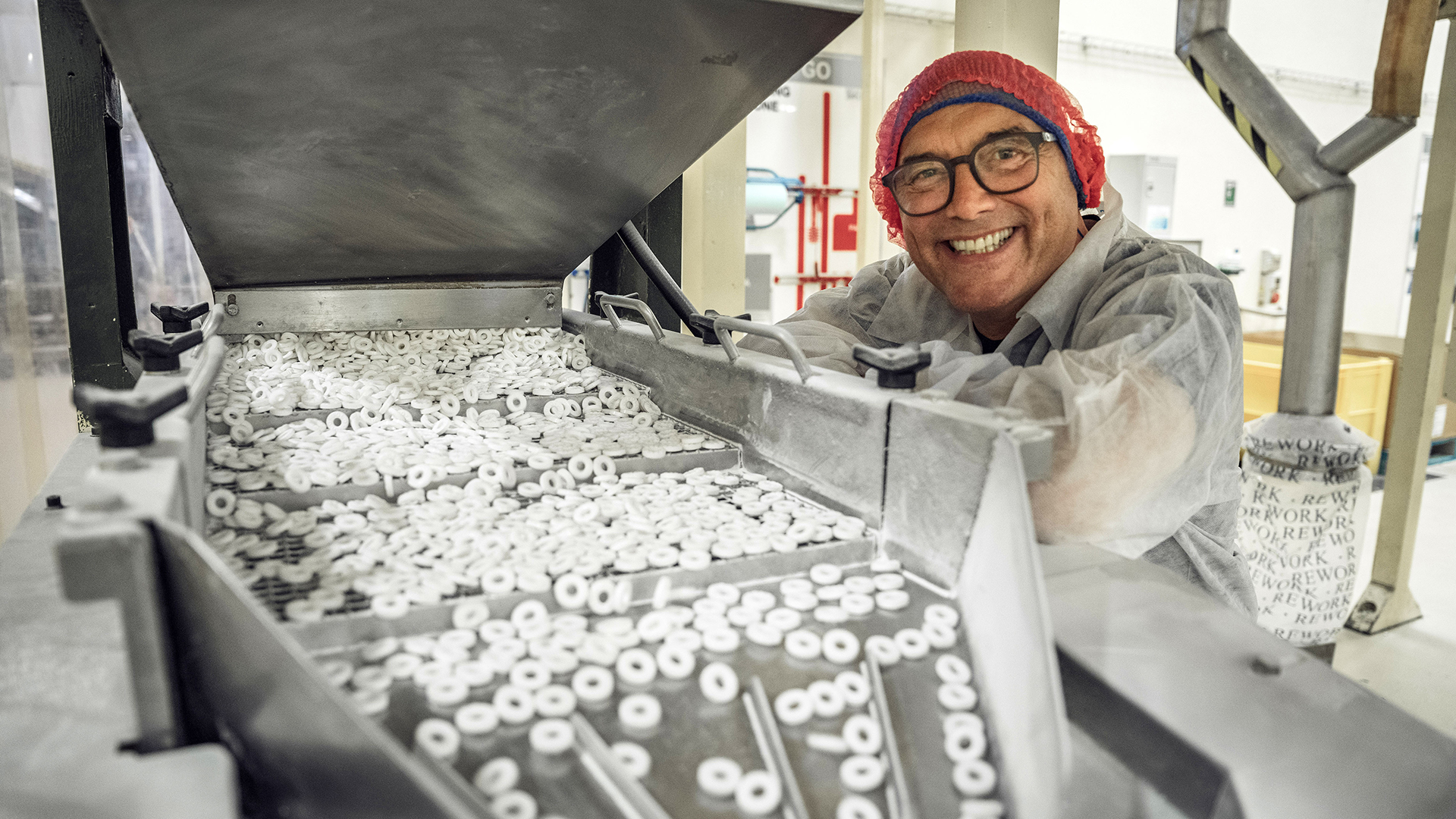 Inside the Factory host Gregg Wallace grinning next to a conveyor belt