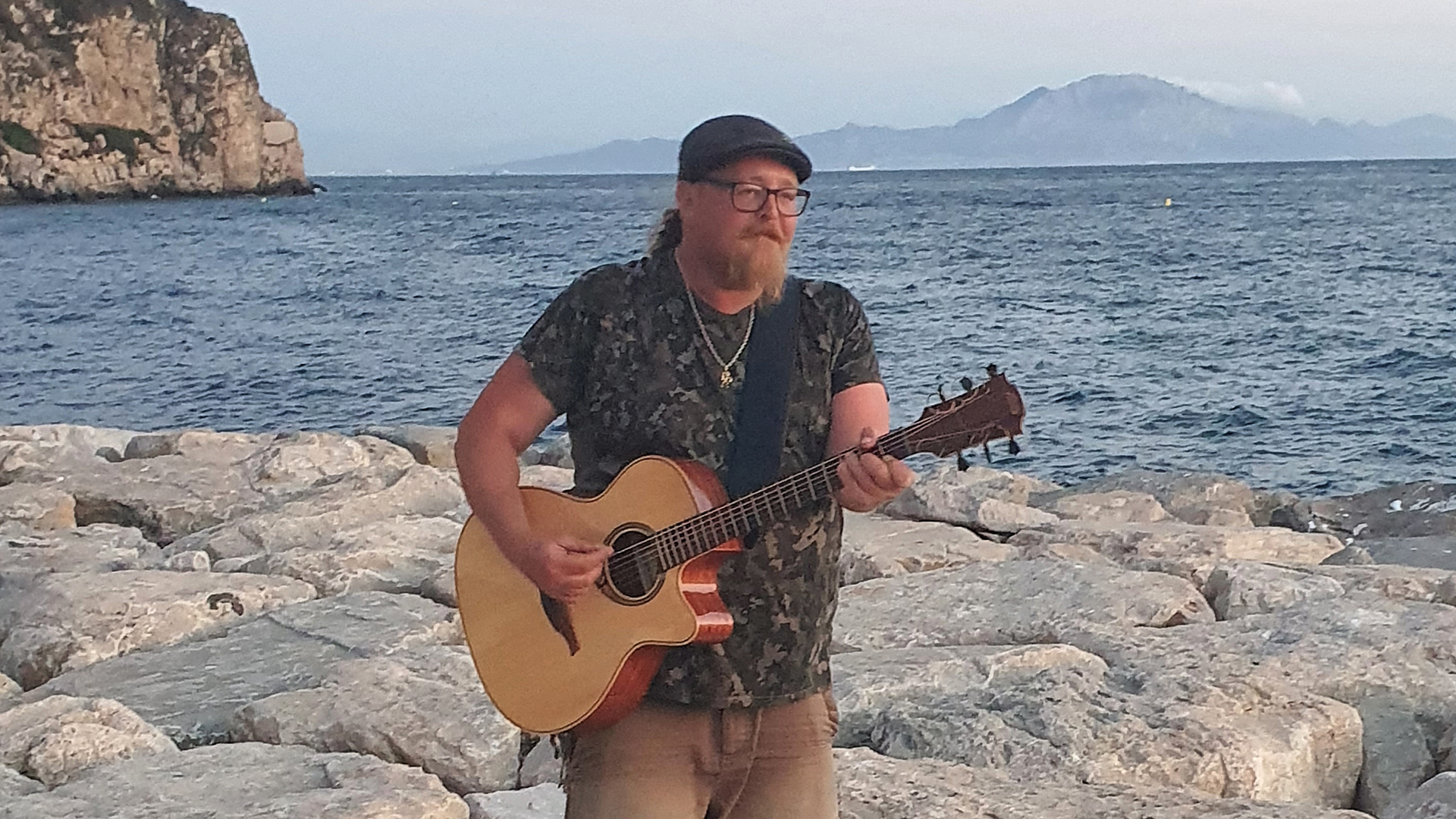 Poorest Tourist, aka Stephen Woodhouse, standing on a beach playing a guitar
