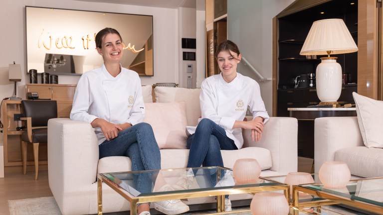 Two young women sit on a sofa in chef's whites with Sacher branding