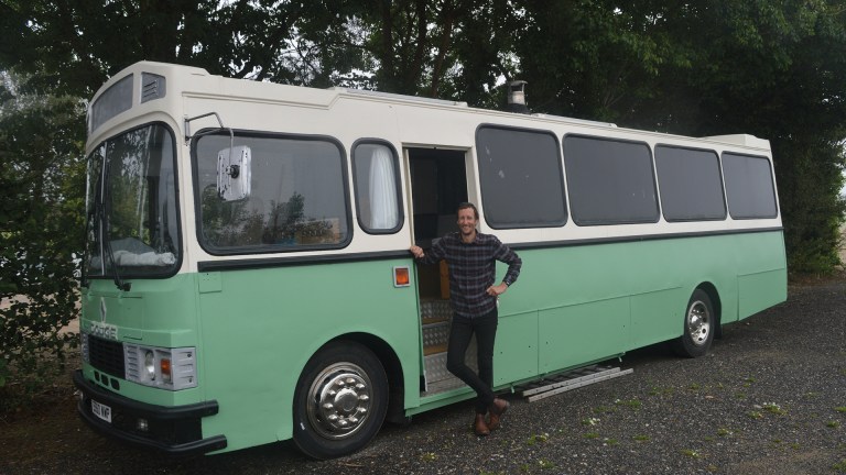 A man stands with his hand on his hip beside a green and white bus