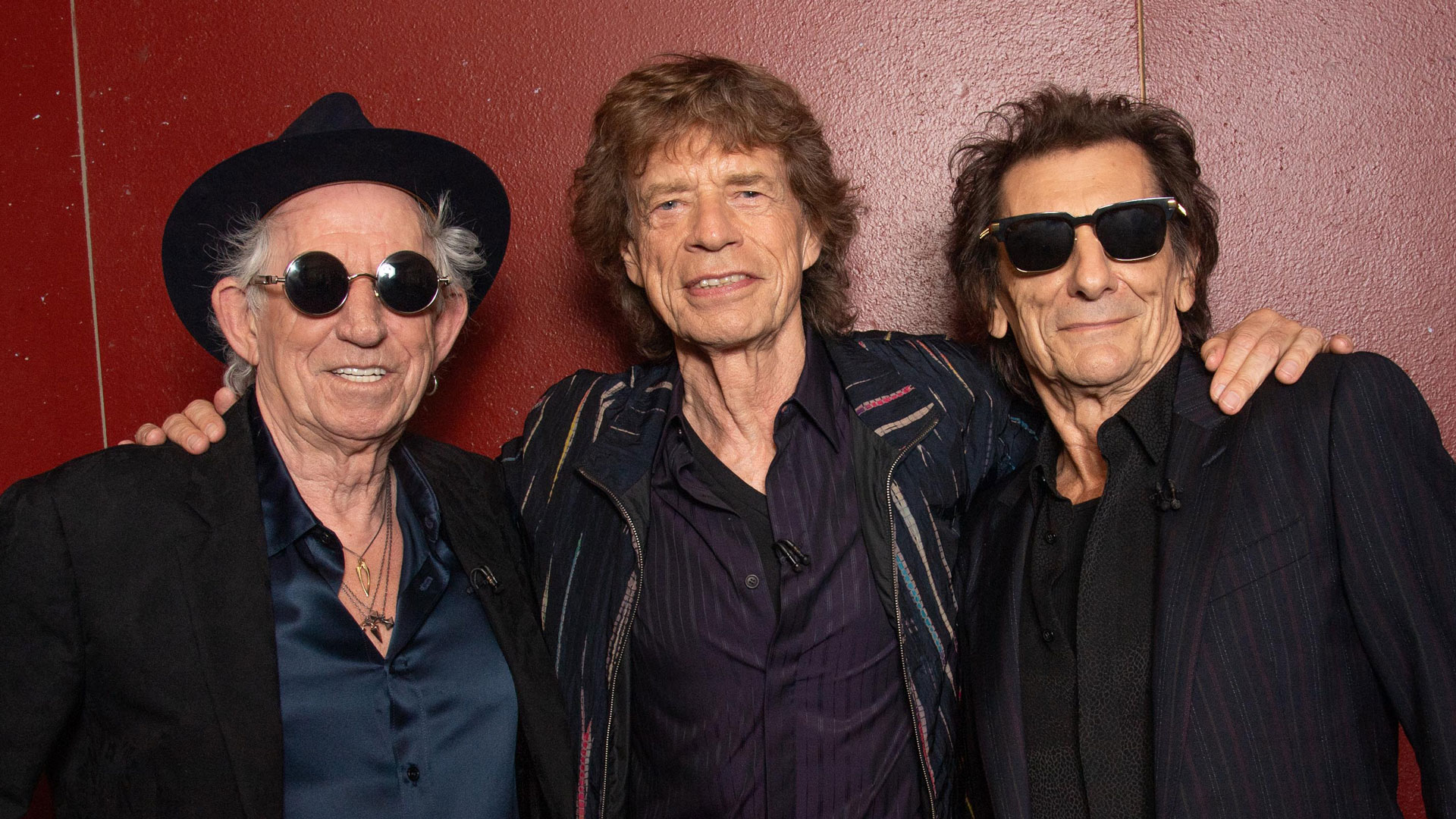 The Rolling Stones: Keith Richards, Mick Jagger and Ronnie Wood in black jackets against a red background