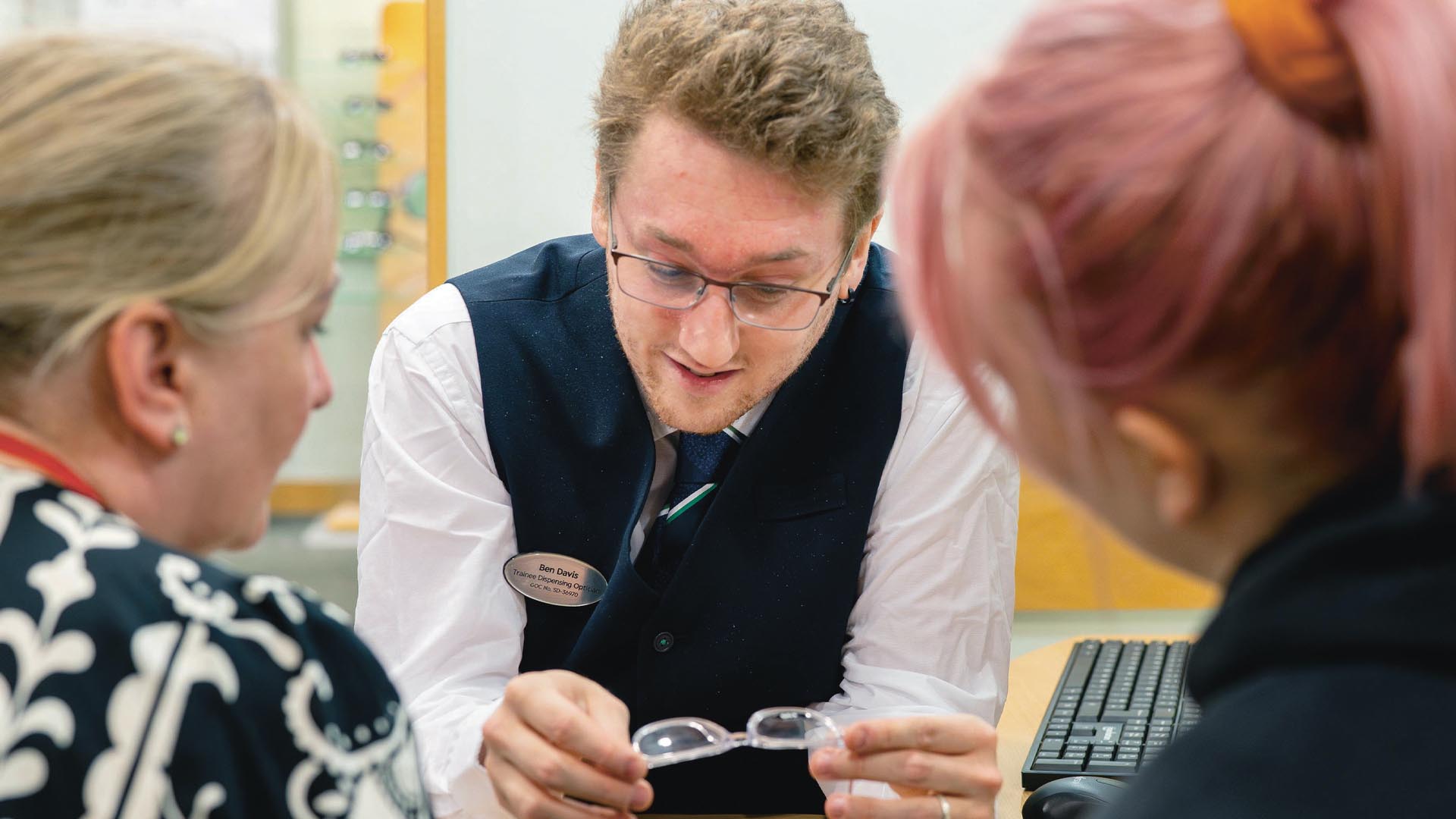 A member of the Specsavers team, male with blonde hair and glasses, looks at a pair of spectacles with his colleagues, both women, one young with pink hair, one older and blonde, wearing a blue and white patterned blouse.