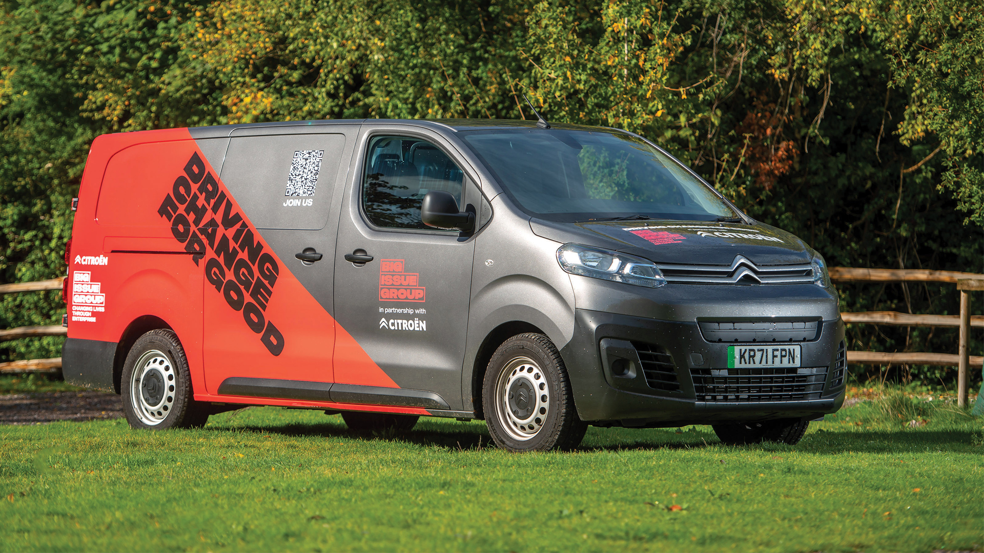 ChatGPT The image shows a Citroën ë-Berlingo van parked on grass with trees in the background, indicating a sunny day. The van is primarily gray with a bold red graphic on the side that reads "DRIVING CHANGE FOR GOOD" along with a QR code and the Big Issue Group and Citroën logos, highlighting their partnership. The registration plate is visible on the front of the van.