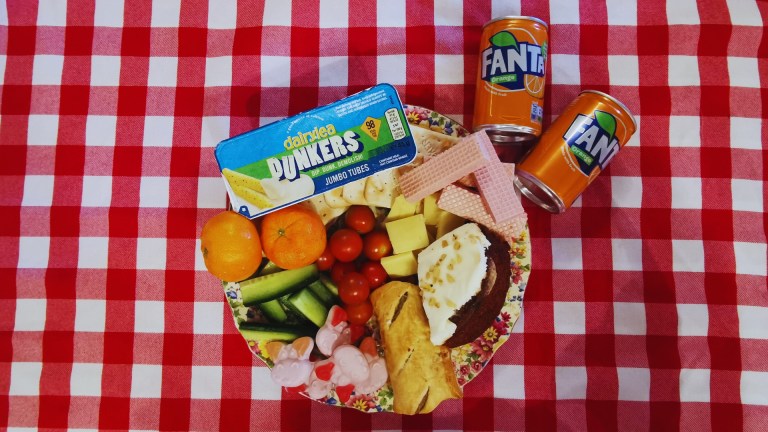 A plate of unmatched food: cake, cucumber, cheese spread and wafers with cans of fanta