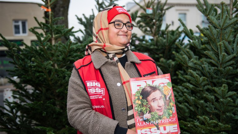 ChatGPT A person wearing a red Big Issue vest and a beige and red striped scarf stands in front of a backdrop of Christmas trees. They are smiling and holding a copy of the Big Issue magazine with a cover featuring Alan Partridge. The individual is wearing glasses and a red beanie hat with the Big Issue logo on it. The outdoor setting suggests it might be cold, and the person is dressed warmly in a jacket with a fleece collar.