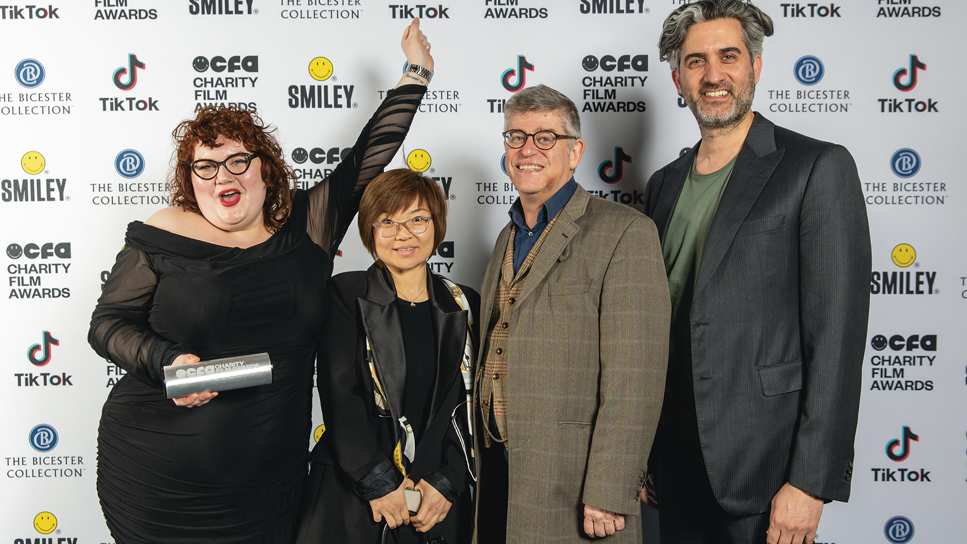 Four individuals from the Welsh Refugee Council pose together at the Charity Film Awards, standing in front of a backdrop featuring sponsor logos. From left to right: a jubilant woman holding an award, raising her arm in celebration; a woman with a short hairstyle, dressed in a black outfit with a printed scarf; a smiling man in a tweed jacket and glasses; and a man with salt-and-pepper hair, wearing a blazer over a casual shirt. All look thrilled to have just won an award.