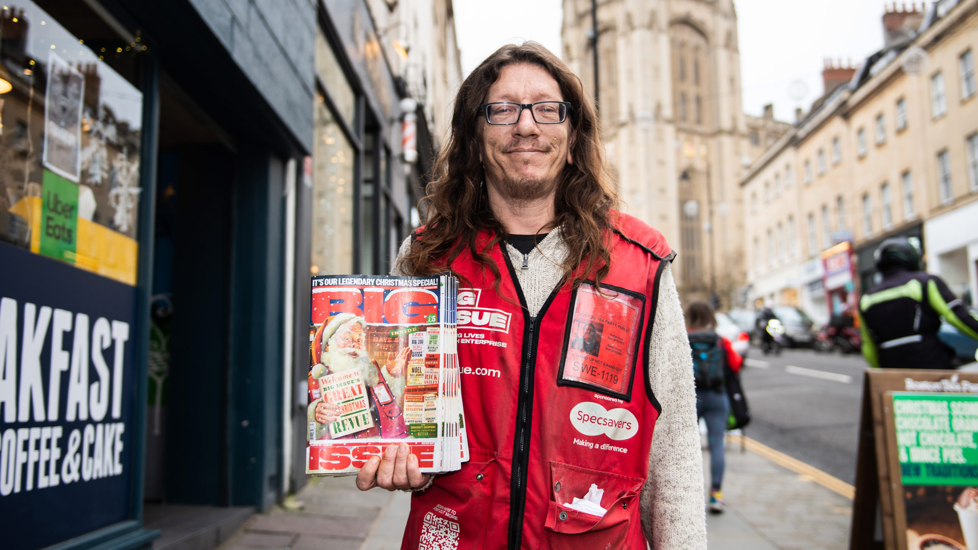 Outdoor image of Jack, a Big Issue vendor, standing on a city street holding a copy of the Big Issue magazine. He's wearing a red vest with the Specsavers logo, signifying their partnership, and glasses. The backdrop features an urban street scene with buildings, pedestrians, and a passing motorcyclist.
