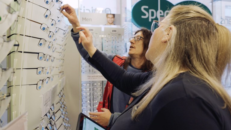 Inside a Specsavers store, a man is selecting glasses from a display rack, assisted by a female store employee. They are both focused on choosing the right pair of frames from a wall of options, under a sign that says 'Men'.