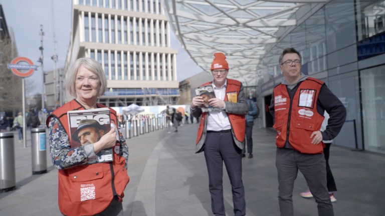 Dame Mary Perkins and John Perkins stand outside King’s Cross station, wearing red Big Issue tabards over their clothes. Dame Mary holds a copy of The Big Issue magazine, and both are sporting red tabards. The bustling background hints at the busy London atmosphere, with a clear sky overhead and modern architecture around. They seem engaged in their role, aiming to understand the experience of magazine vendors.