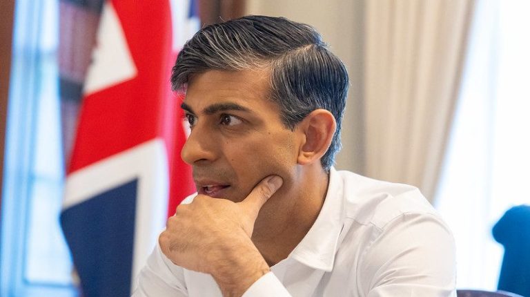Prime minister Rishi Sunak in a white shirt sat at his desk in front of a Union Jack flag