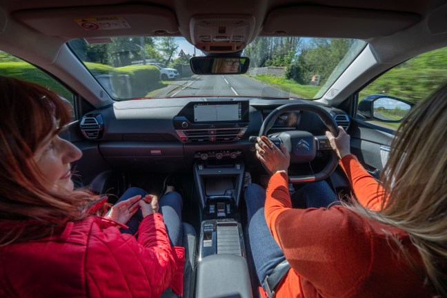 This image shows two women inside a car during a daytime drive. The driver, wearing an orange sweater, is focused on the road while operating the car's controls. The passenger, in a red jacket, sits relaxed with her hands folded in her lap. The car's interior features a modern dashboard with a digital display for navigation. The view through the windshield shows a quiet, suburban road lined with greenery under a clear sky. The scene captures a moment of travel, possibly engaging in a conversation, in a comfortably appointed vehicle.