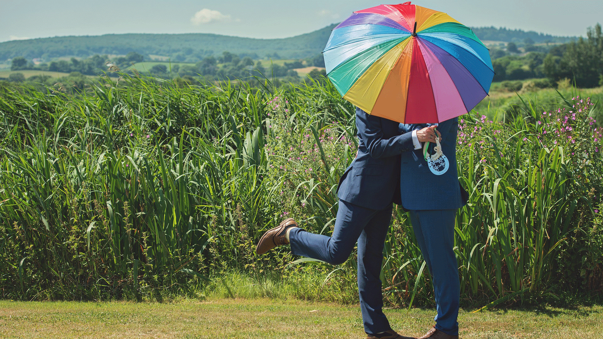 Queer love: Two men standing under a rainbow umbrella in front of a field