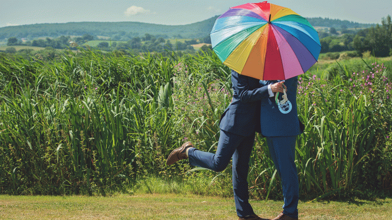 Queer love: Two men standing under a rainbow umbrella in front of a field