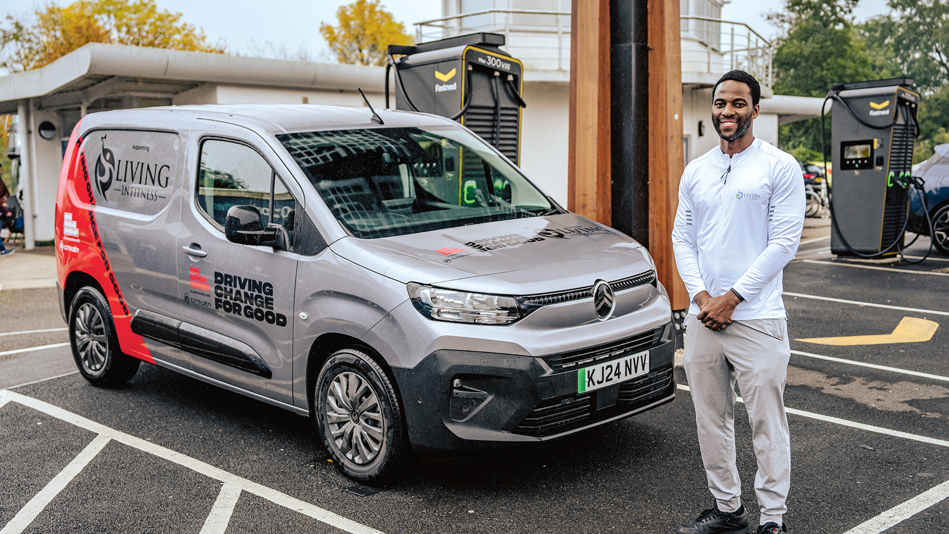 A man wearing a white sports top and light trousers stands smiling next to a grey Citroën ë-Berlingo van with “Living in Fitness” and “Driving Change for Good” branding. The van is parked at an electric vehicle charging station with a Fastned charger in the background. The setting is an outdoor charging area with a covered structure and trees with autumn foliage.