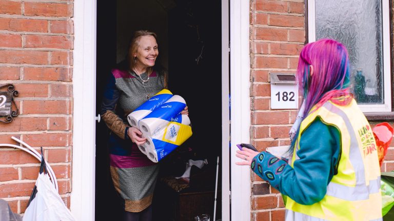 A person in a door receives a package of toilet roll from a person in a high-vis jacket