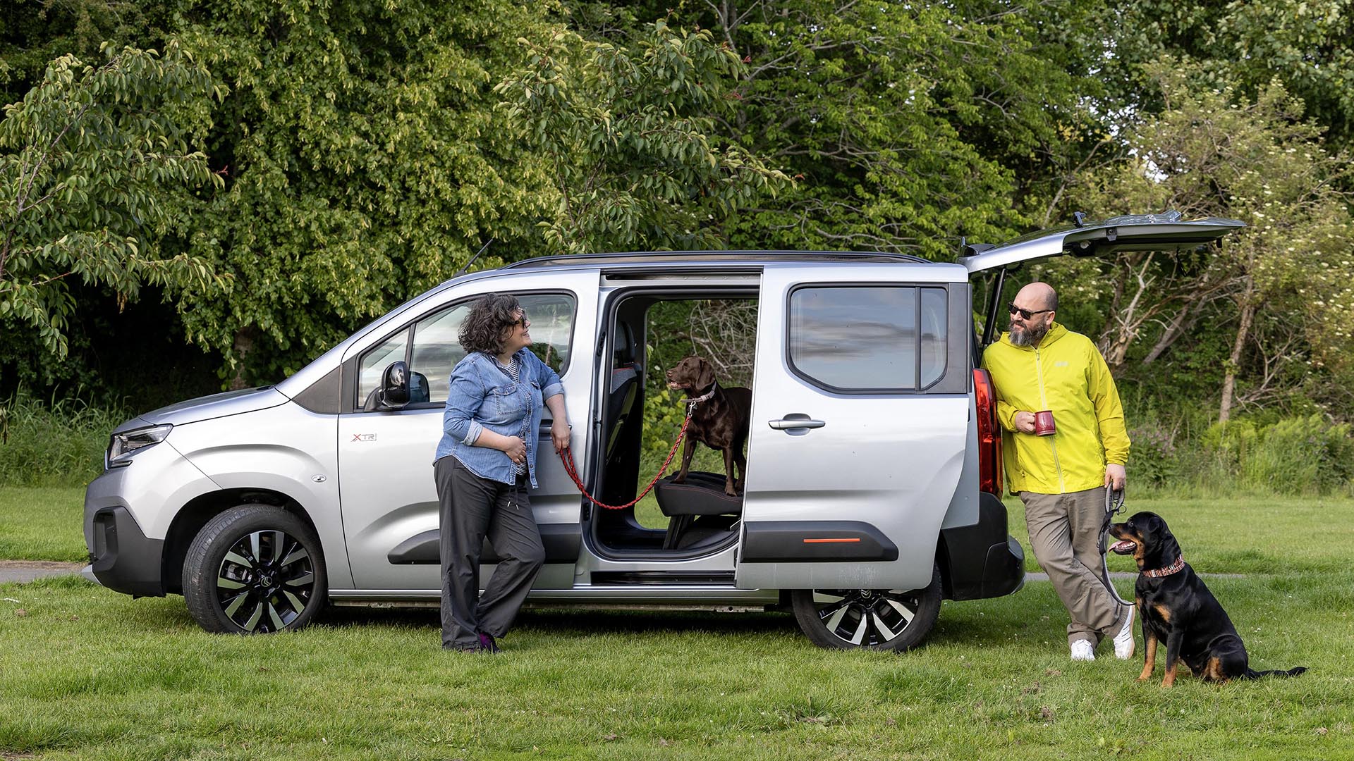 Couple with dogs standing beside silver Citroën ë-Berlingo with sliding doors open showing spacious interior