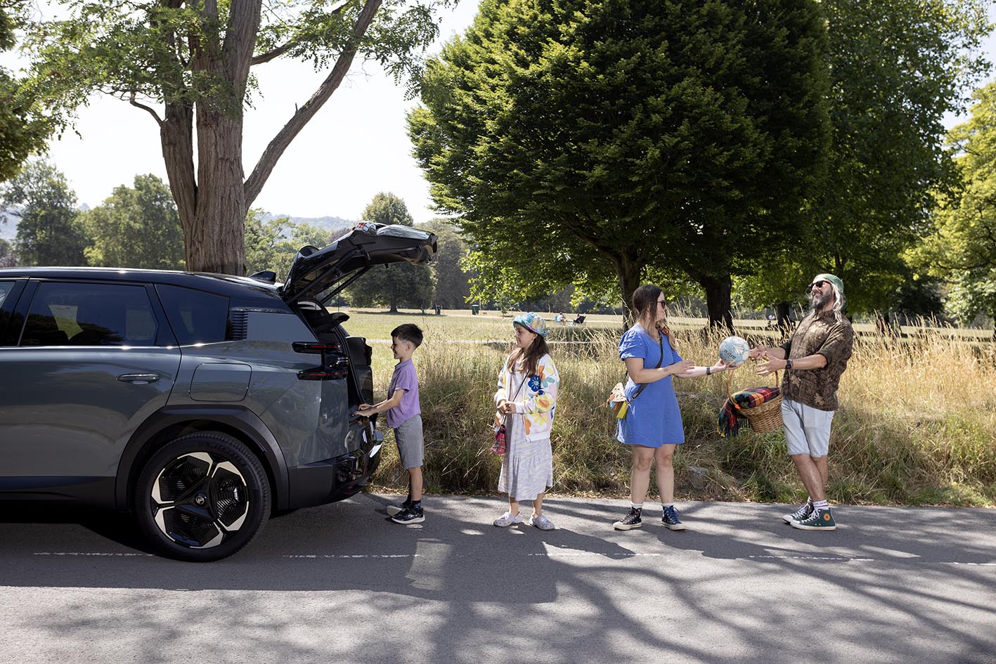 A family of four unpacking picnic supplies from the open boot of a grey Citroën ë-C5 Aircross parked on a country lane, with green fields and trees in the background on a sunny day.