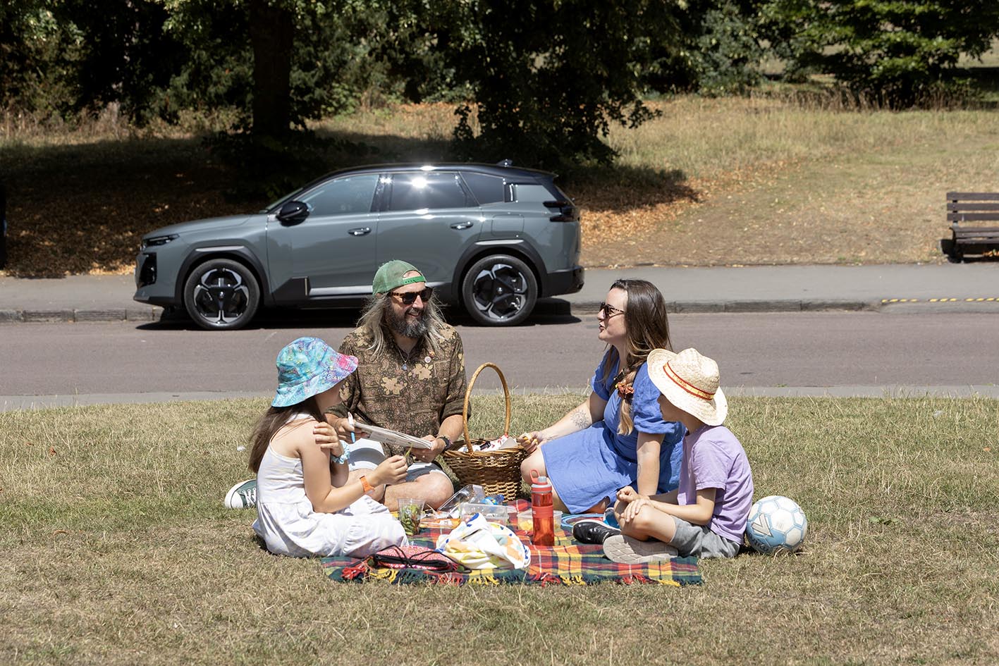 A family enjoying a picnic on a tartan blanket on grass parkland, with their grey Citroën ë-C5 Aircross parked nearby under the shade of large trees on a sunny summer day.