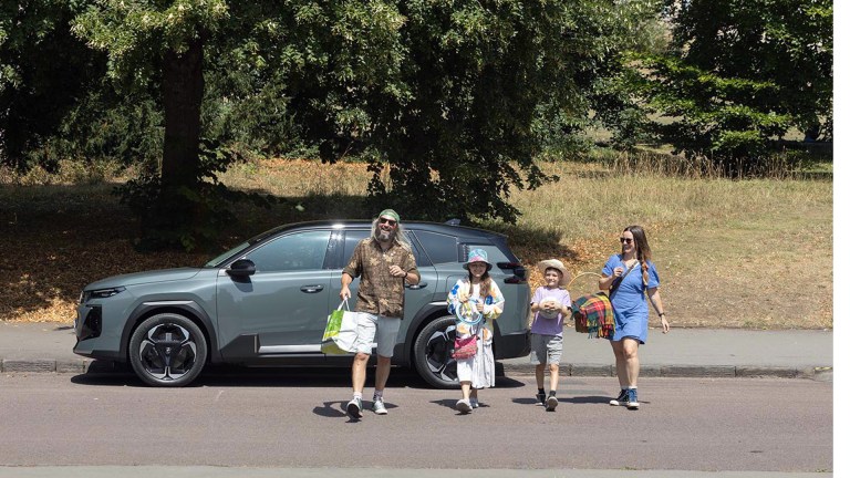 A family of four with picnic basket and sports equipment standing beside their grey Citroën ë-C5 Aircross, parked under trees in a countryside setting with golden grass fields visible behind them.
