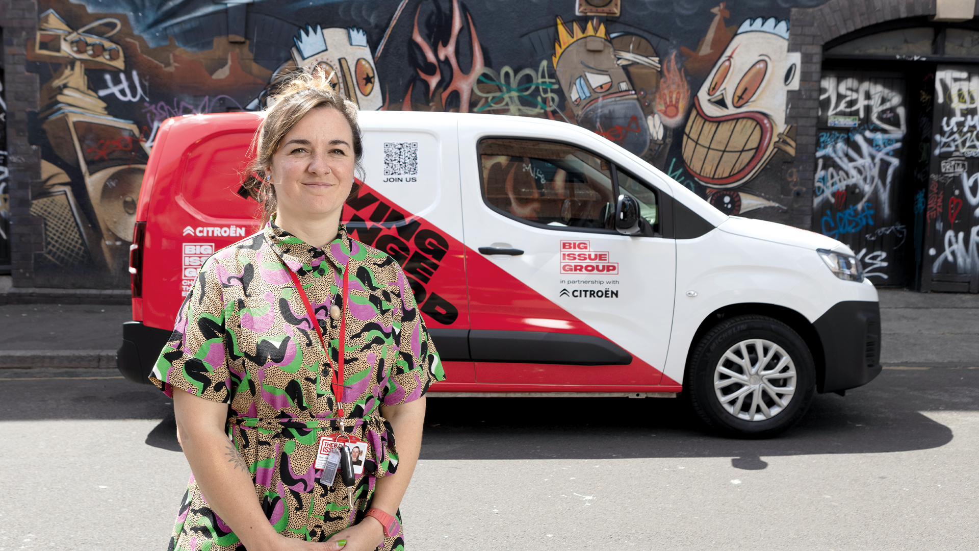 Big Issue frontline manager Hattie Greenyer standing with a Big Issue magazine in front of the Citroën ë-Berlingo Van displaying 'Driving Change for Good' branding, with colourful street art mural in background